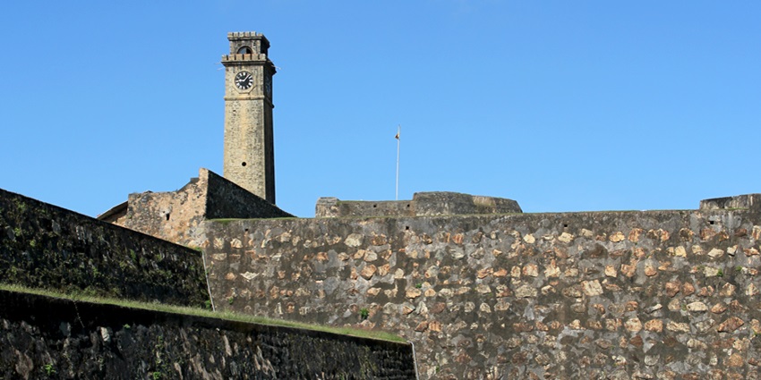 Historic Galle Fort with a prominent clock tower, surrounded by greenery and blue sky.