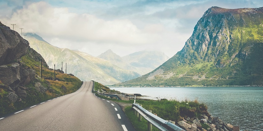 A view of a narrow strip of road with majestic mountains and an ocean in the background.
