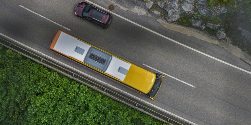 An aerial view of a yellow bus and a white car in the middle of the road around trees.