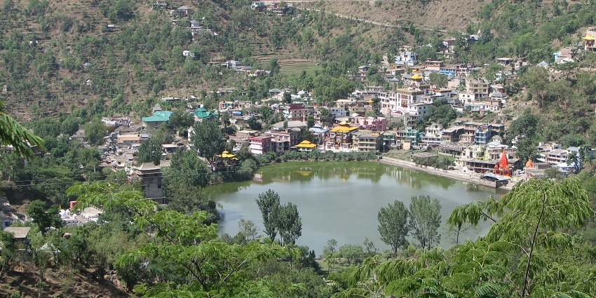 : A breathtaking view of the Kamru Nag Lake surrounded by mountains and houses all around.