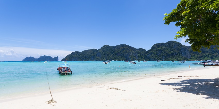 A picture of a beach in Pattaya with a speedboat sailing in the ocean and a hilly island in the background