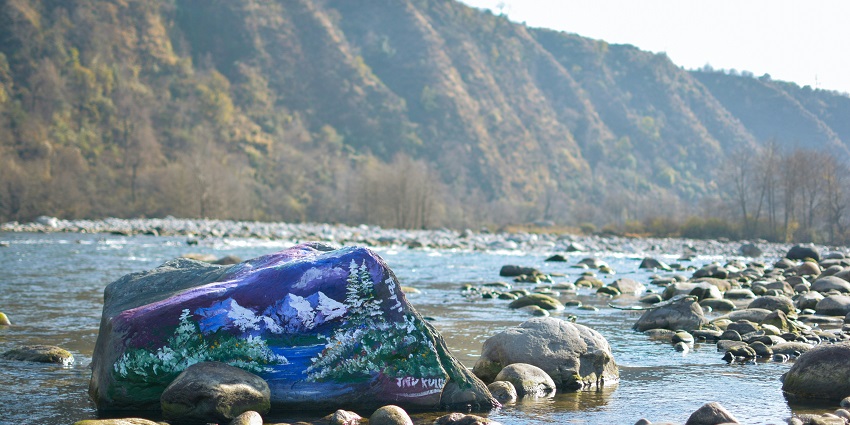 Image of a large blue rock sits in a flowing river, surrounded by lush greenery.
