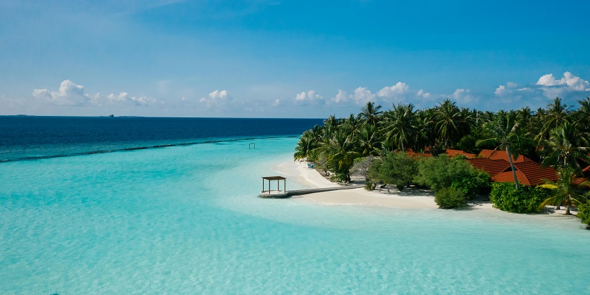 A drone shot of a beach which has crystal-clear water and is surrounded by green palm trees.