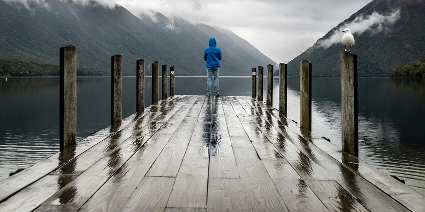 Lakeside view with brown Wooden Dock and a boy standing, surrounded by green mountains.
