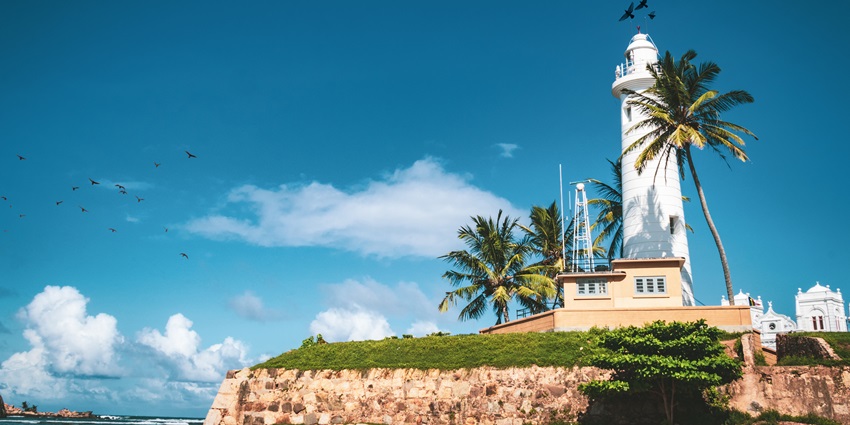 White lighthouse at Galle, Sri Lanka, standing tall against a bright blue sky and palm trees.