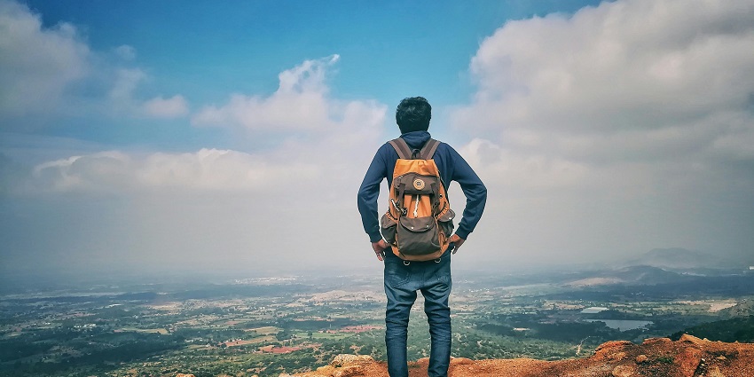 A man in a blue shirt and jeans stands on a cliff, gazing at a town under a blue sky.