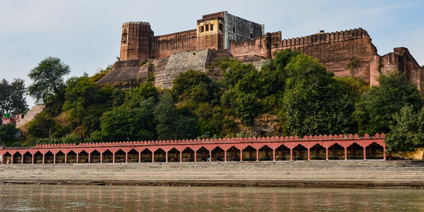 A riverside picture of a majestic fort with tall outer walls and a bushy area in front