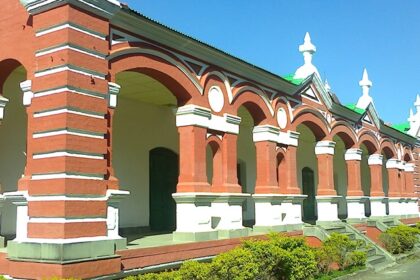 A frontal view of a red and white concrete building with lush greenery during the day.