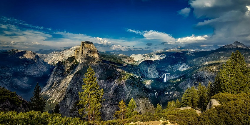 Panoramic mountain view with towering peaks, lush valleys, and a clear blue sky.