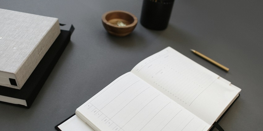 View of three diaries, a yellow pencil & a black pen stand kept on a grey-coloured table.