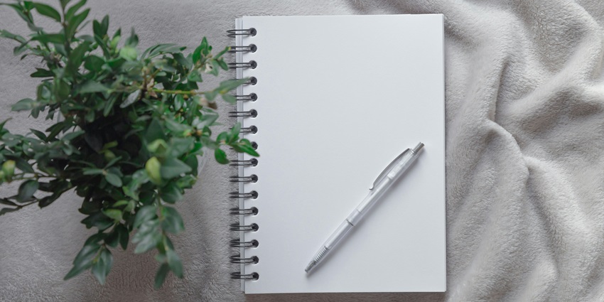 An image of a spiral notebook with a pen and a plant kept on a white towel on a table.