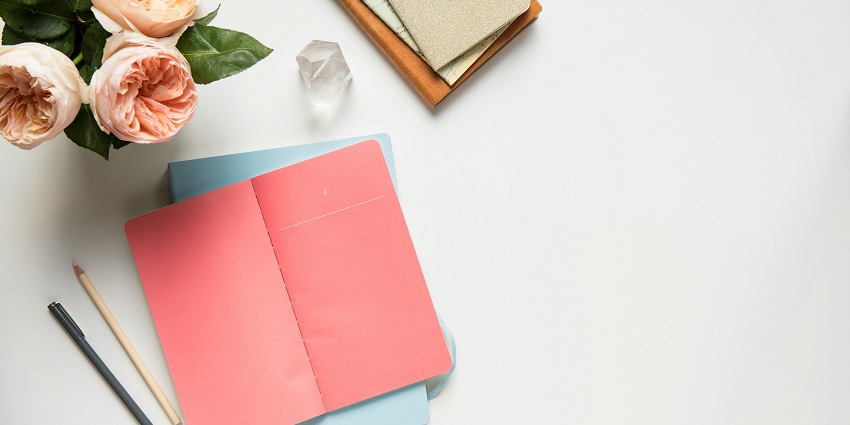 A picture of multiple notebooks, a pen, a pencil, and flowers kept on a white table.