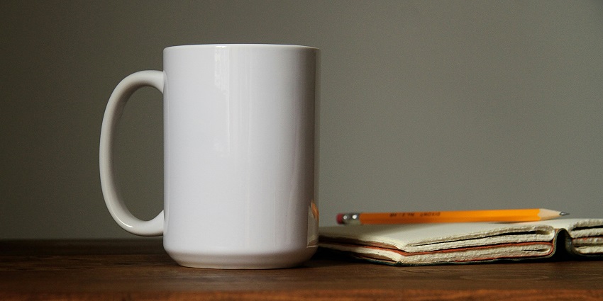 A picture of a white ceramic mug, with a notebook and pencil, kept on a wooden table.