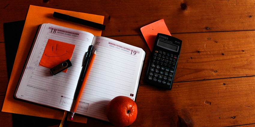 View of a black calculator kept on a table with a notebook, orange and pencil on the side.