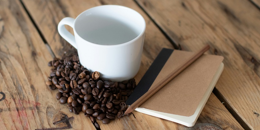 Picture of a cup of coffee kept on a wooden table with a pencil and notebook by its side.