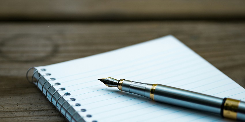 View of a spiral notebook with ruled pages & a fountain pen kept on a wooden brown table.