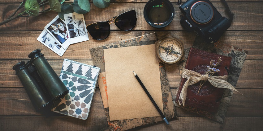 An image of a pencil, paper, camera, binoculars, compass, and sunglasses kept on a table.