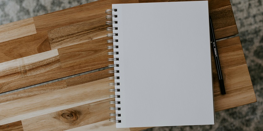 A picture of a pencil and a spiral notebook with white pages lying on a wooden table.