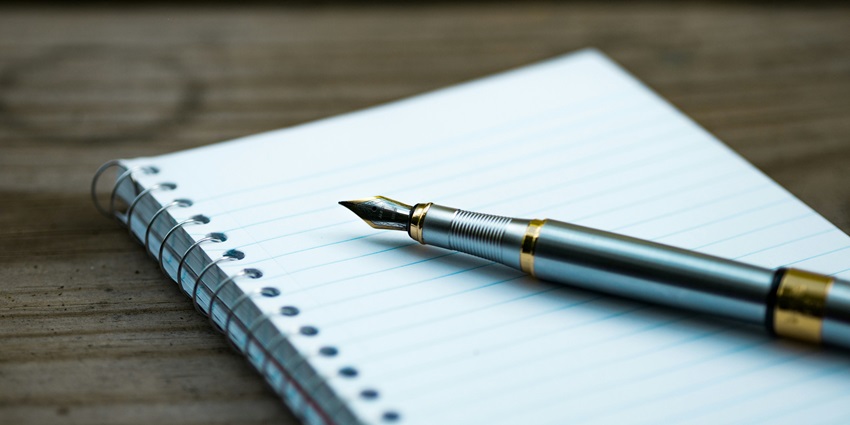 View of a notebook with spiral binding and a fountain pen on it lying on a wooden table.