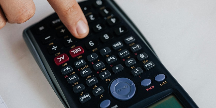 A close-up view of a finger using a black calculator on plain white surface with numbers.