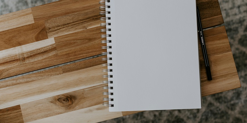 An aerial shot of an open white notebook with a pen on the side sitting on a table.