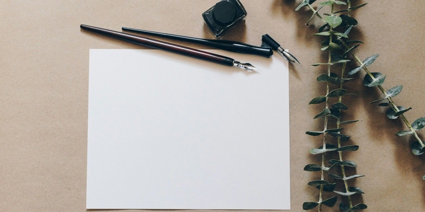 A view of a white blank paper with ink pens on the side with a green leaf on the table.