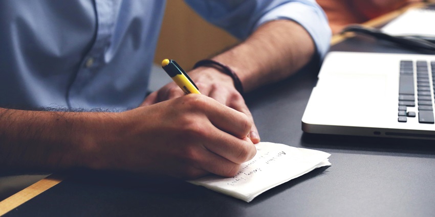 A focused view of a man’s hand holding a yellow and black pen with a notebook in front.