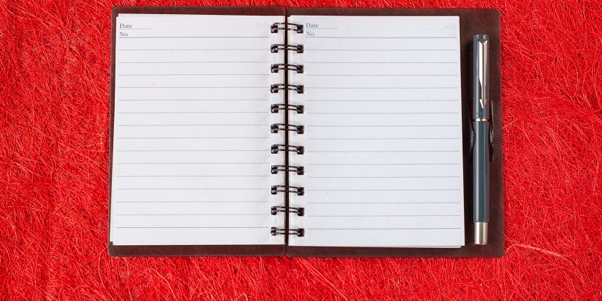 View of an open black notebook with white pages with a pen on the side sitting on a table.