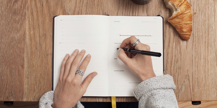 A person writing on a white notebook with a black pen and a cup of coffee on the side.