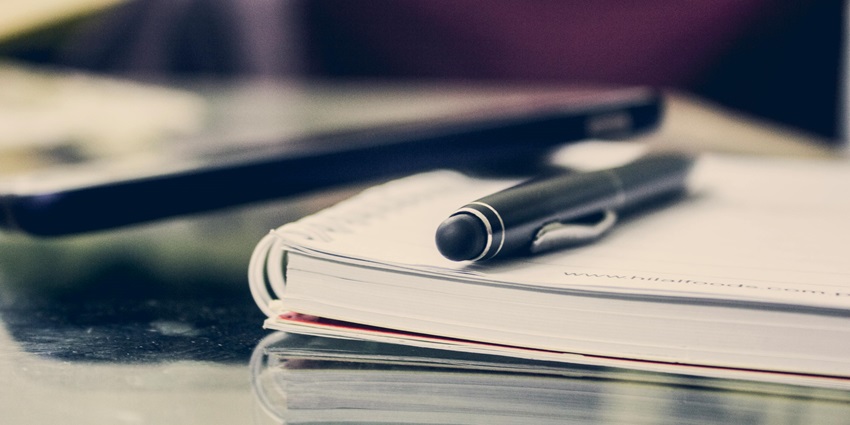 A focused view of a notebook with a black pen on it kept on a table during the day.