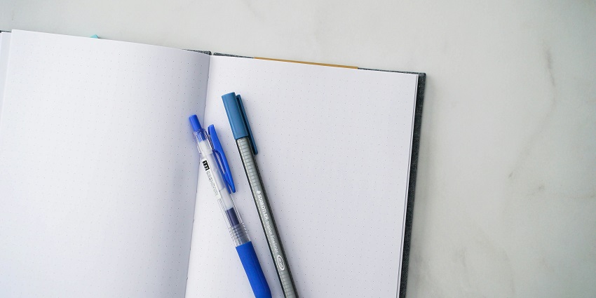An aerial shot of a notebook with white blank pages and a few different pens on top of it.