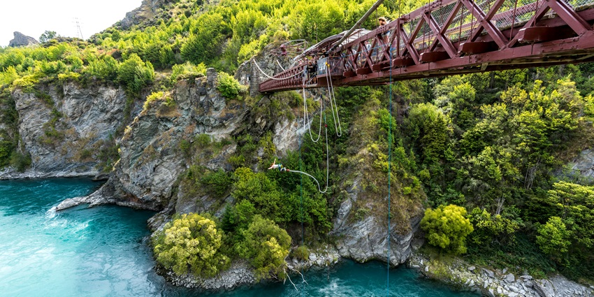 A vibrant red metal bridge spanning a lush green gorge, surrounded by dense forest.