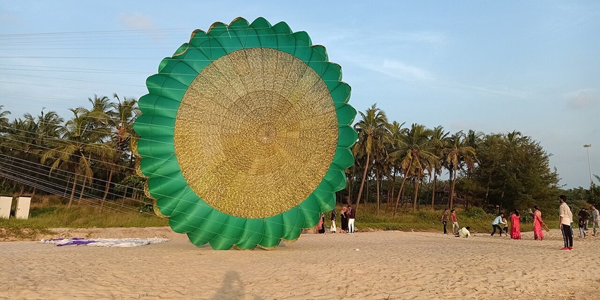 Circular kite soars above a sandy beach as spectators line the shore at Bekal