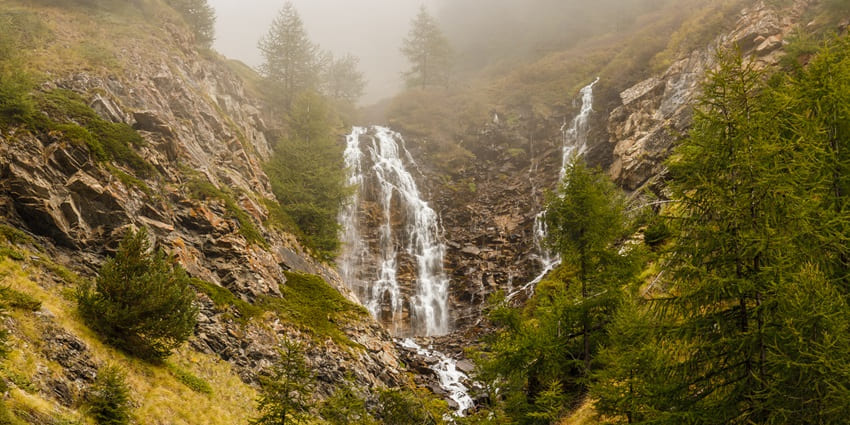 A picture of a majestic waterfall with a small stream, cascading down a lush green hill