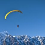 A scenic view of a person soaring high above in the air over the snow-capped peaks.