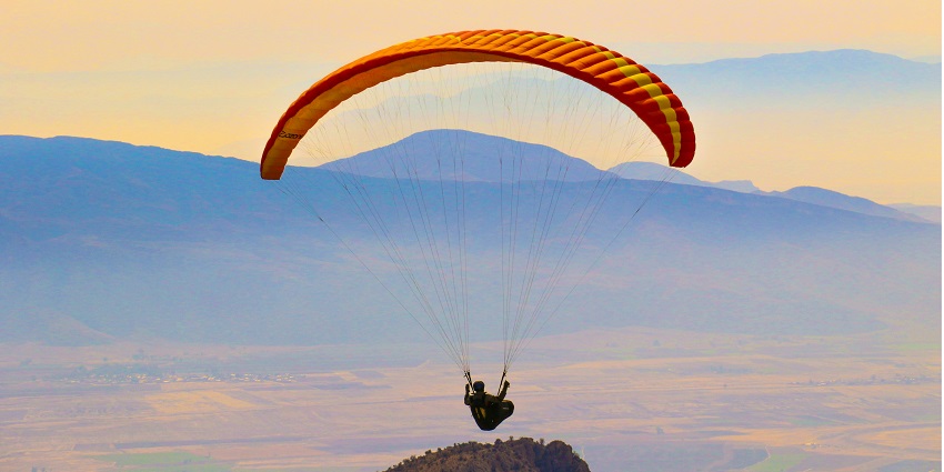 A person flying high up in the air above the peaks with a colourful backdrop of sky.