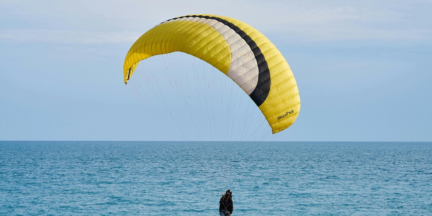 Person with a yellow and white gliding parachute soaring near the sea, with waves below.