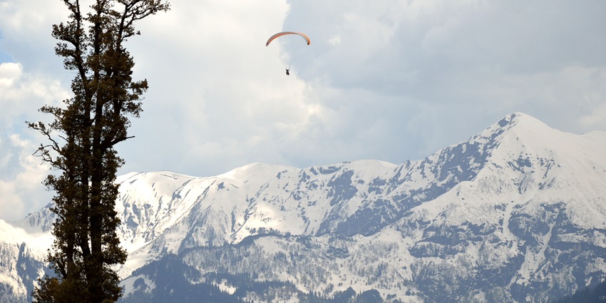 Image of a paraglider soaring over lush green hills in Gulaba, Kullu, under a clear blue sky.
