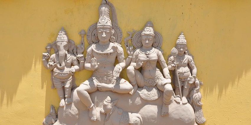 A picture of a sculpture of Hindu deities made with clay, placed against a yellow wall