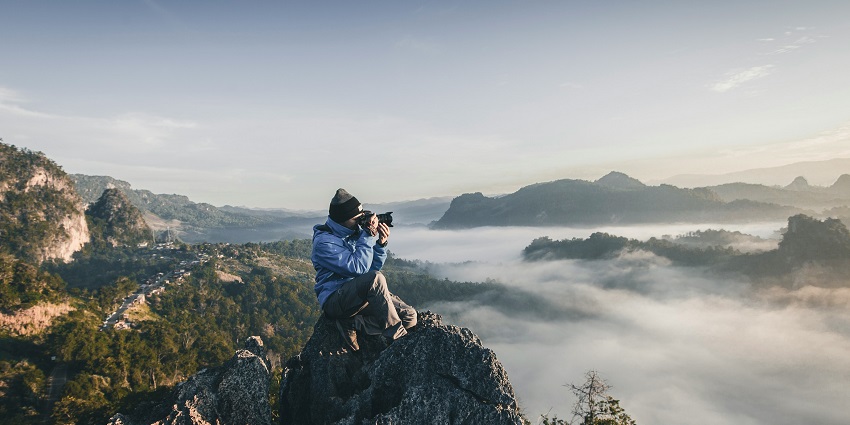 A picture of a man sitting on the top of a rocky mountain and taking photographs.