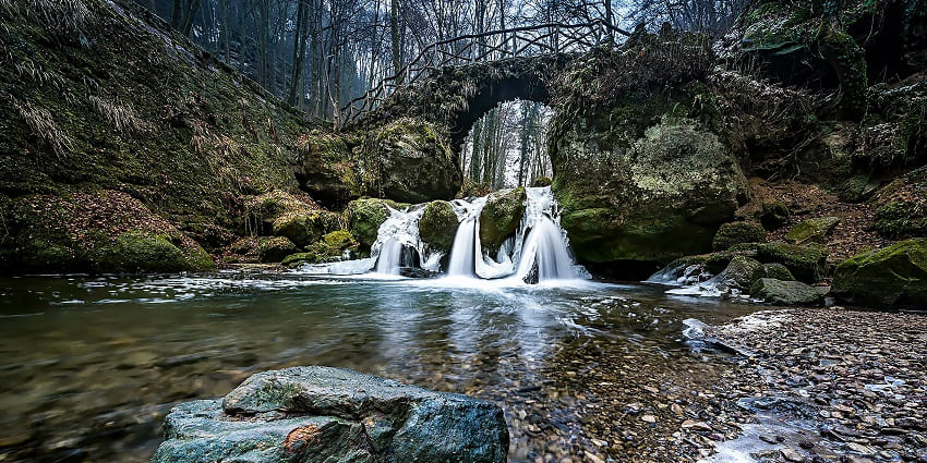 A peaceful river meanders over stones, bordered by lush foliage and a clear, sunny sky.