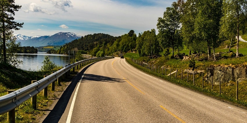 Serene view of road beside the lake, surrounded by lush greenery under clear blue sky.