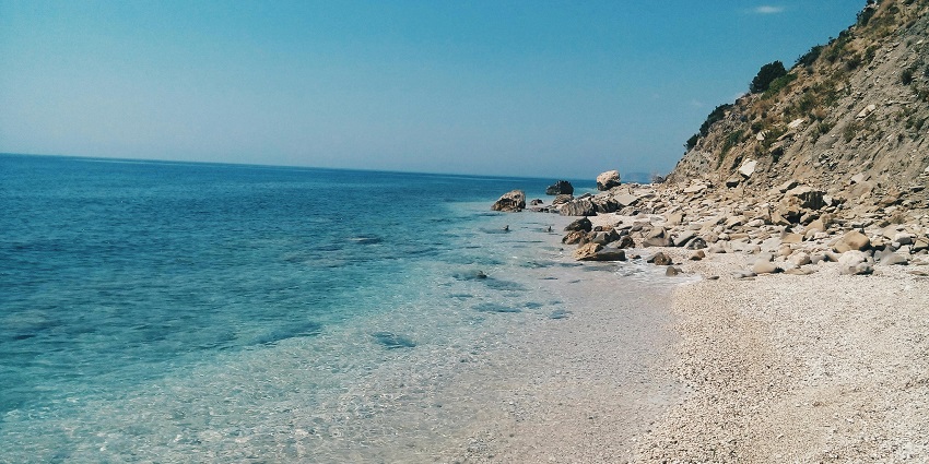 Large grey rock formation by a vibrant blue beach under a bright daytime sky.