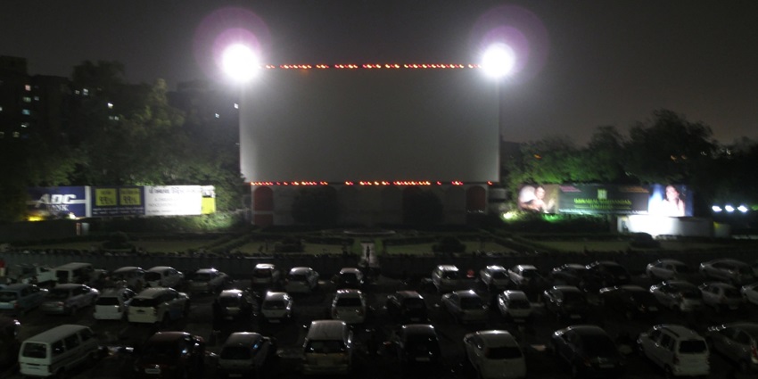 A picture of a drive-in cinema screen with numerous cars parked in front to enjoy movies