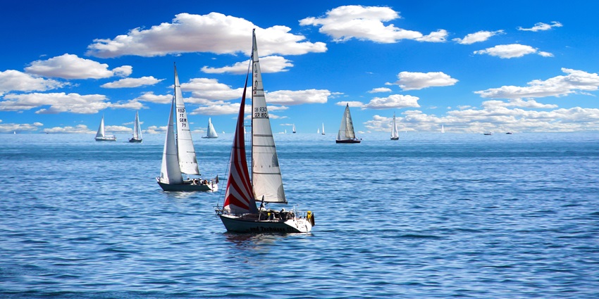 A group of boats sailing through the ocean on a bright day with white clouds in the sky