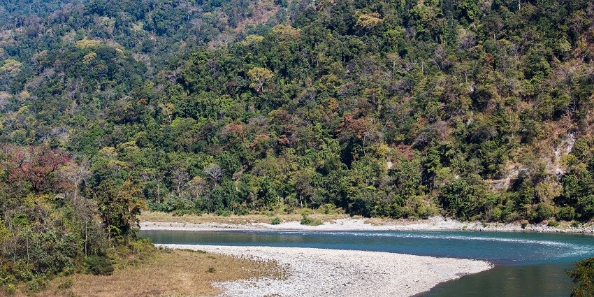 Rocky shores alongside a turquoise stream at the feet of lush forested hills