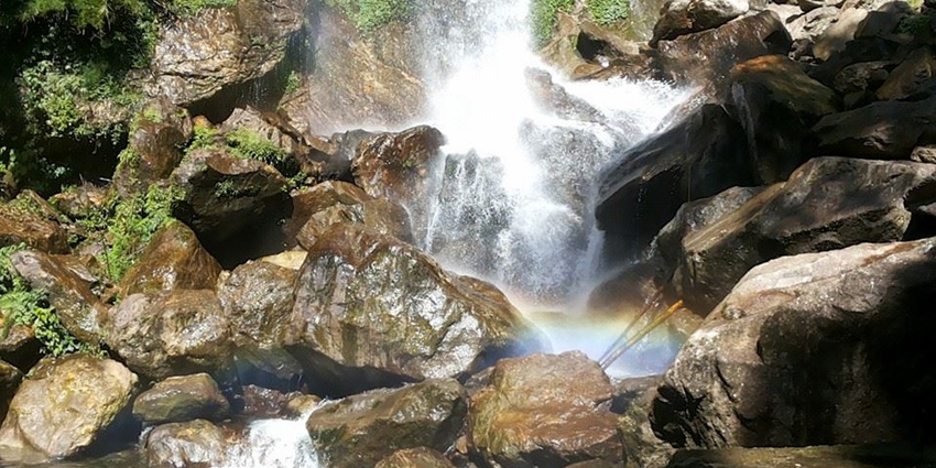 A majestic picture of a waterfall in Sikkim with a rainbow forming around the stream, amidst the rocky terrain