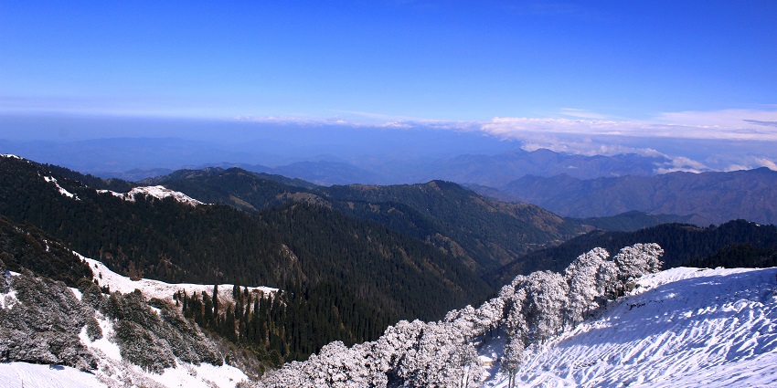 A view of the Shikari Devi Wildlife Sanctuary with snow-covered peaks and trees.