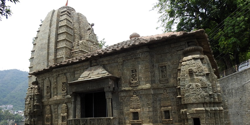 View of a holy place in Himachal Pradesh with lush green trees & mountains around it.