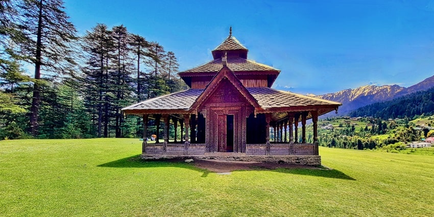 View of a temple surrounded by lush greenery and snow-clad peaks in the background.